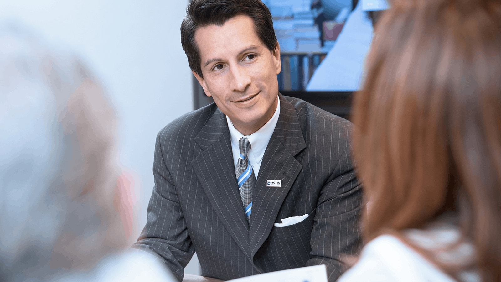 A man in a pinstripe suit with a name badge sits at a table, smiling and listening attentively to two people whose backs are visible in the foreground.