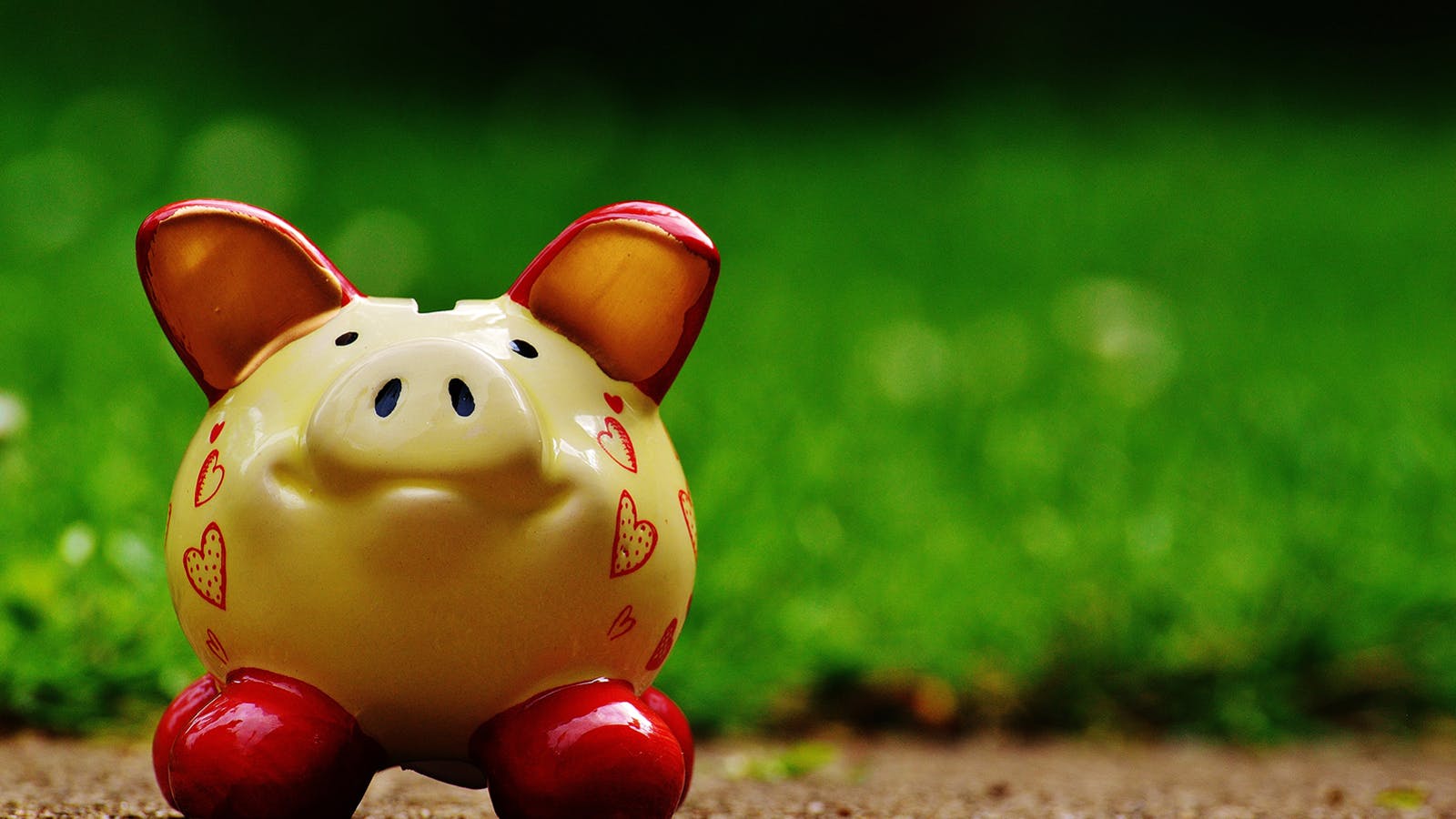 A yellow piggy bank decorated with red hearts and red feet stands on a brown surface, with a blurred green background of grass.