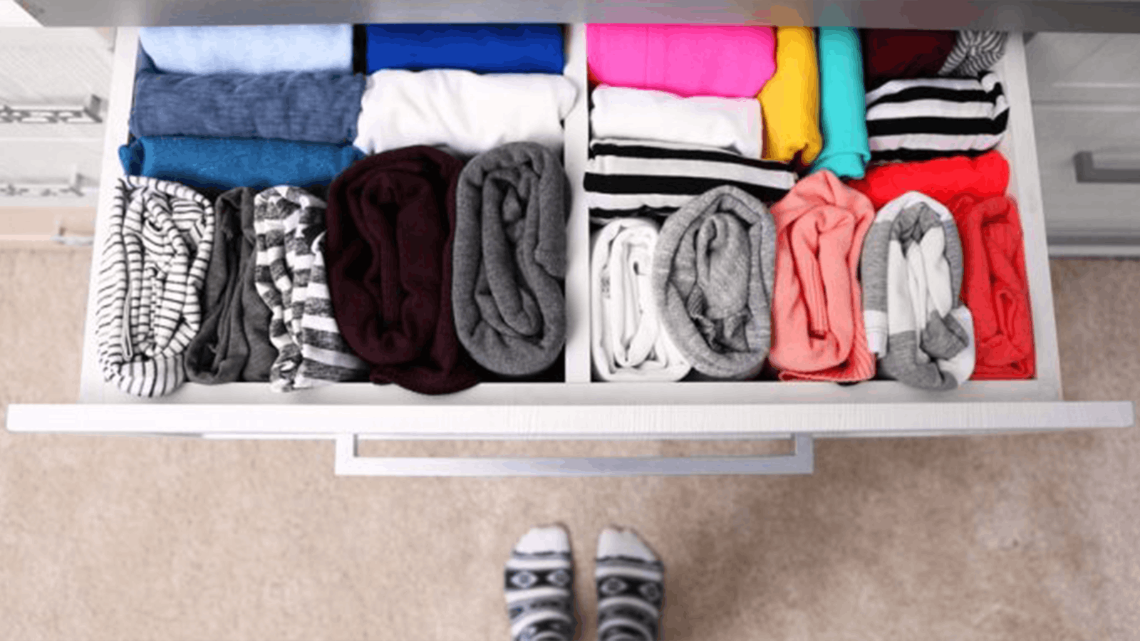 An organized drawer with neatly rolled and folded clothes in various colors and patterns, viewed from above, with a pair of socked feet visible at the bottom of the image.