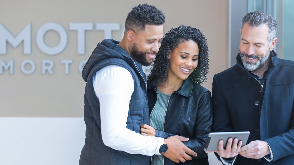 A smiling couple stands close together, looking at a tablet held by a man with a beard. They appear to be in a modern office, possibly discussing something with a professional.