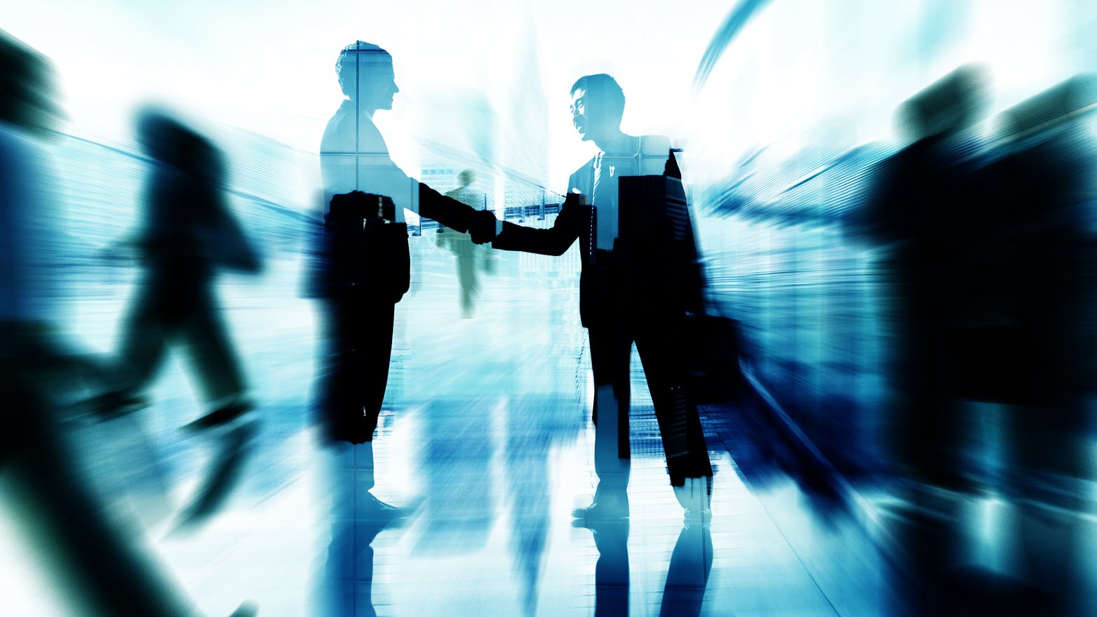Two businesspeople in suits shake hands in a bright, modern office space. Their silhouettes are highlighted, with blurred figures walking around them, creating a sense of motion and busy activity.