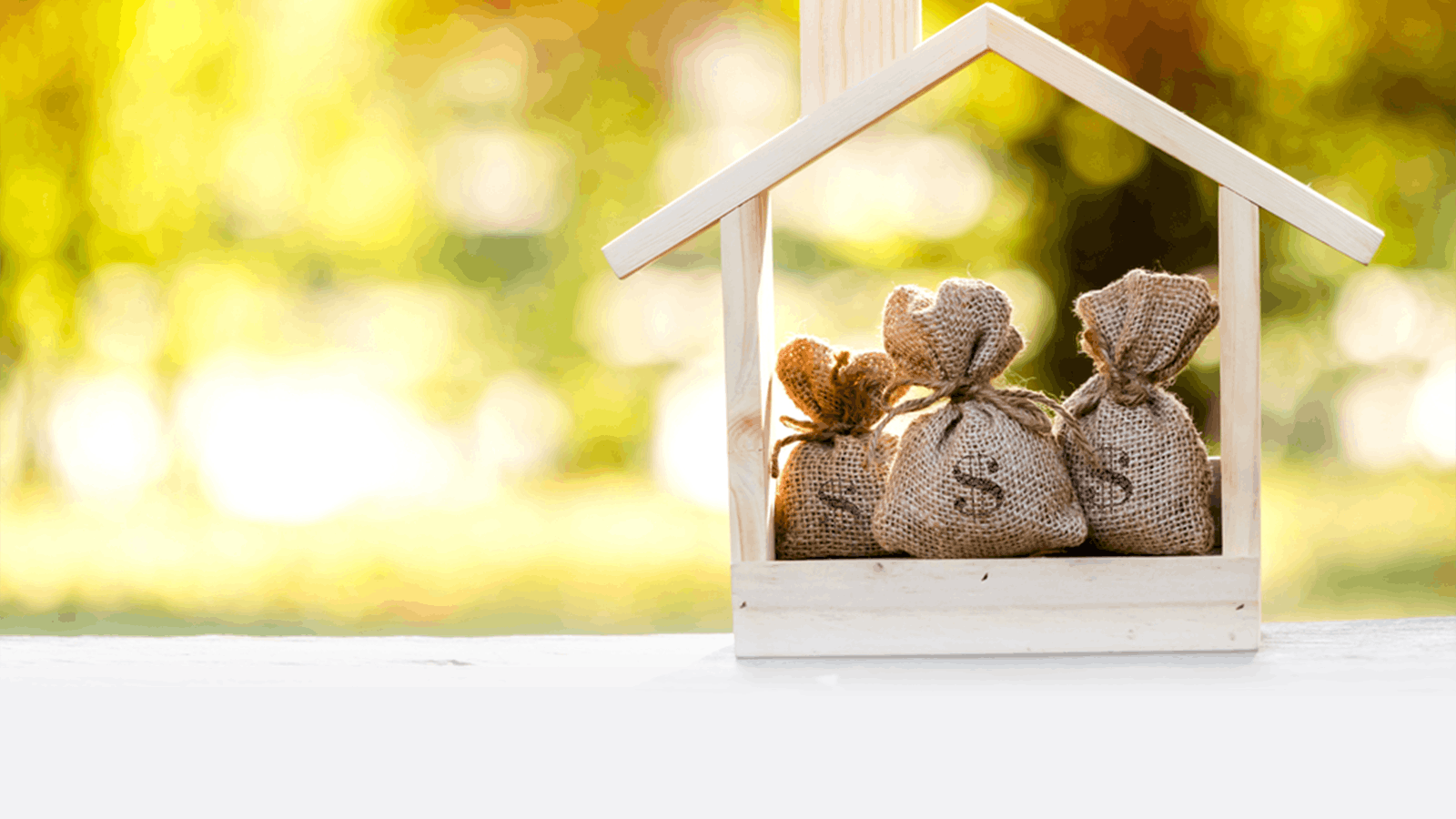 Three small burlap bags with dollar signs sit inside a wooden house-shaped frame, symbolizing savings or investment for a home, against a blurred natural background with sunlight.