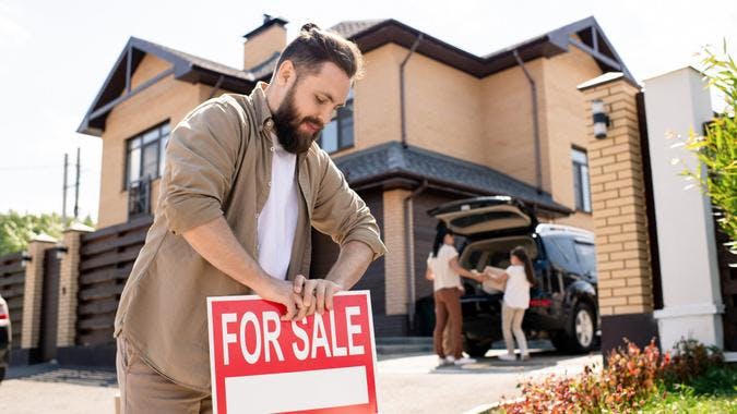 A man places a For Sale sign in front of a modern house, while two people load items into an SUV in the driveway.
