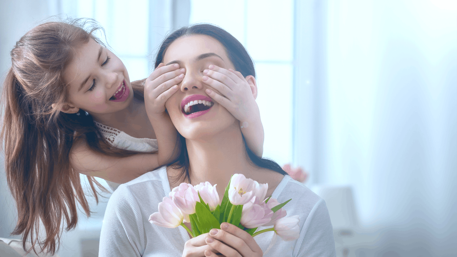 A smiling young woman holding pink tulips is playfully surprised by a little girl who covers her eyes from behind. Both look happy in a bright, softly lit room.
