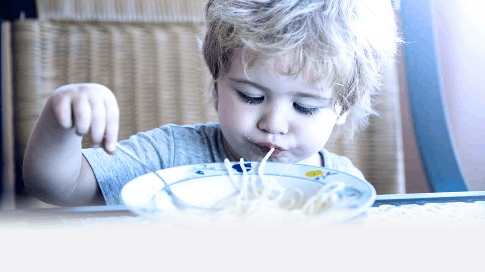 A young child with blonde hair is eating spaghetti from a plate, using their hands and mouth to pull up the noodles while sitting at a table.