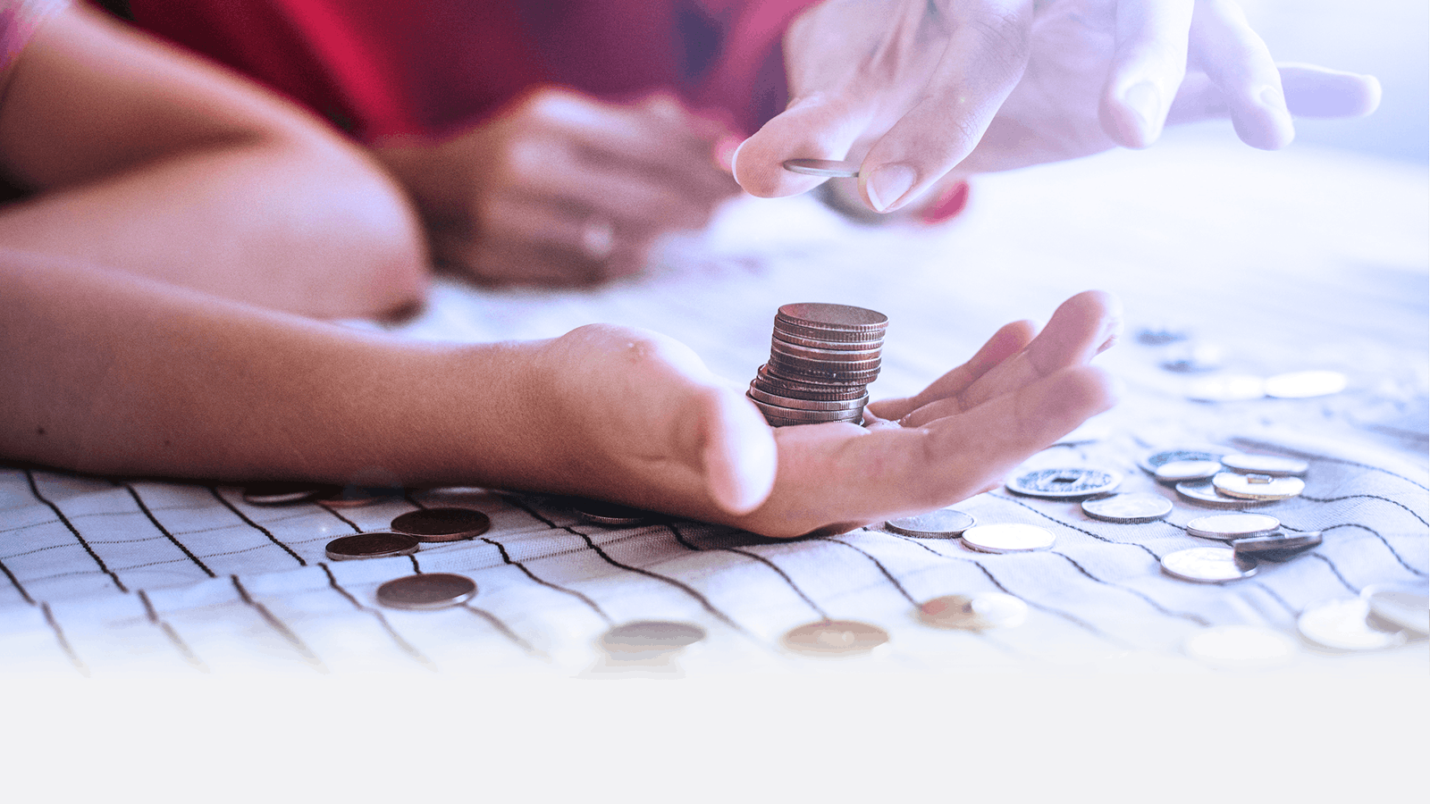 Two hands exchange coins over a table covered with scattered coins and a checkered cloth. One hand holds a stack of coins while the other hand reaches to add another coin.