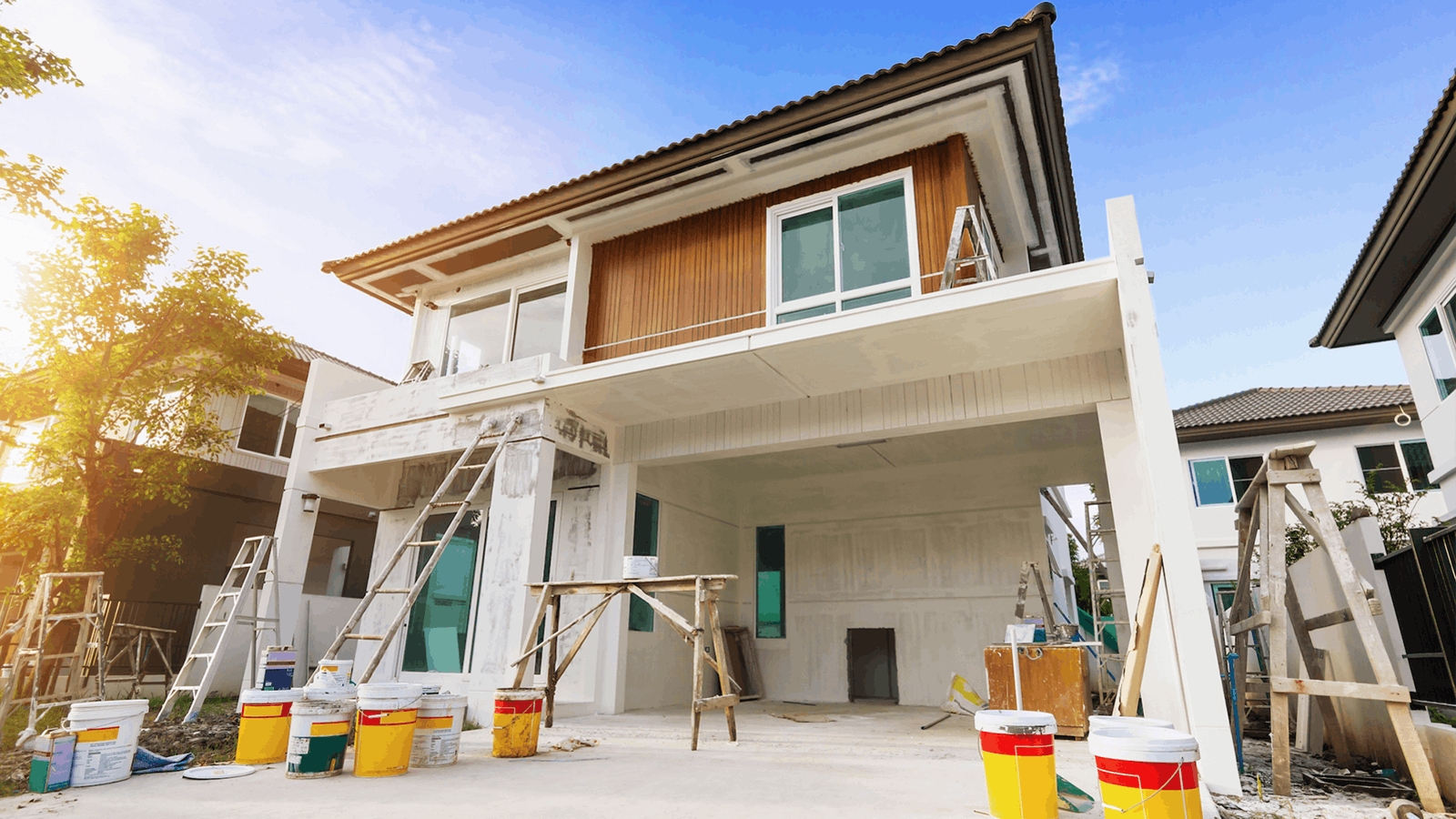 A two-story house under renovation with scaffolding, ladders, and paint buckets in front. The exterior walls are partially painted, and large windows are visible. Trees and another house are in the background.