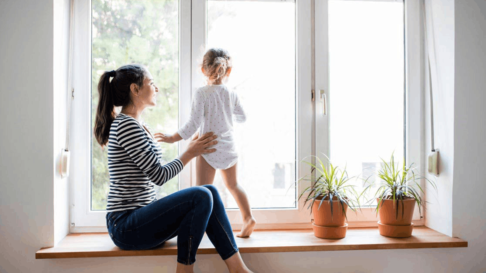 A woman sits on a wide windowsill, smiling at a young child standing beside her and looking out the window. Two potted plants are placed on the sill near them. Natural light fills the room.