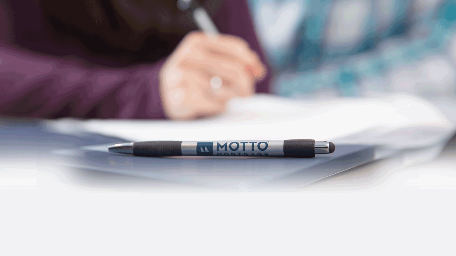 A close-up of a pen with the logo Motto Mortgage printed on it, lying on a table. In the blurred background, a person is writing on papers.