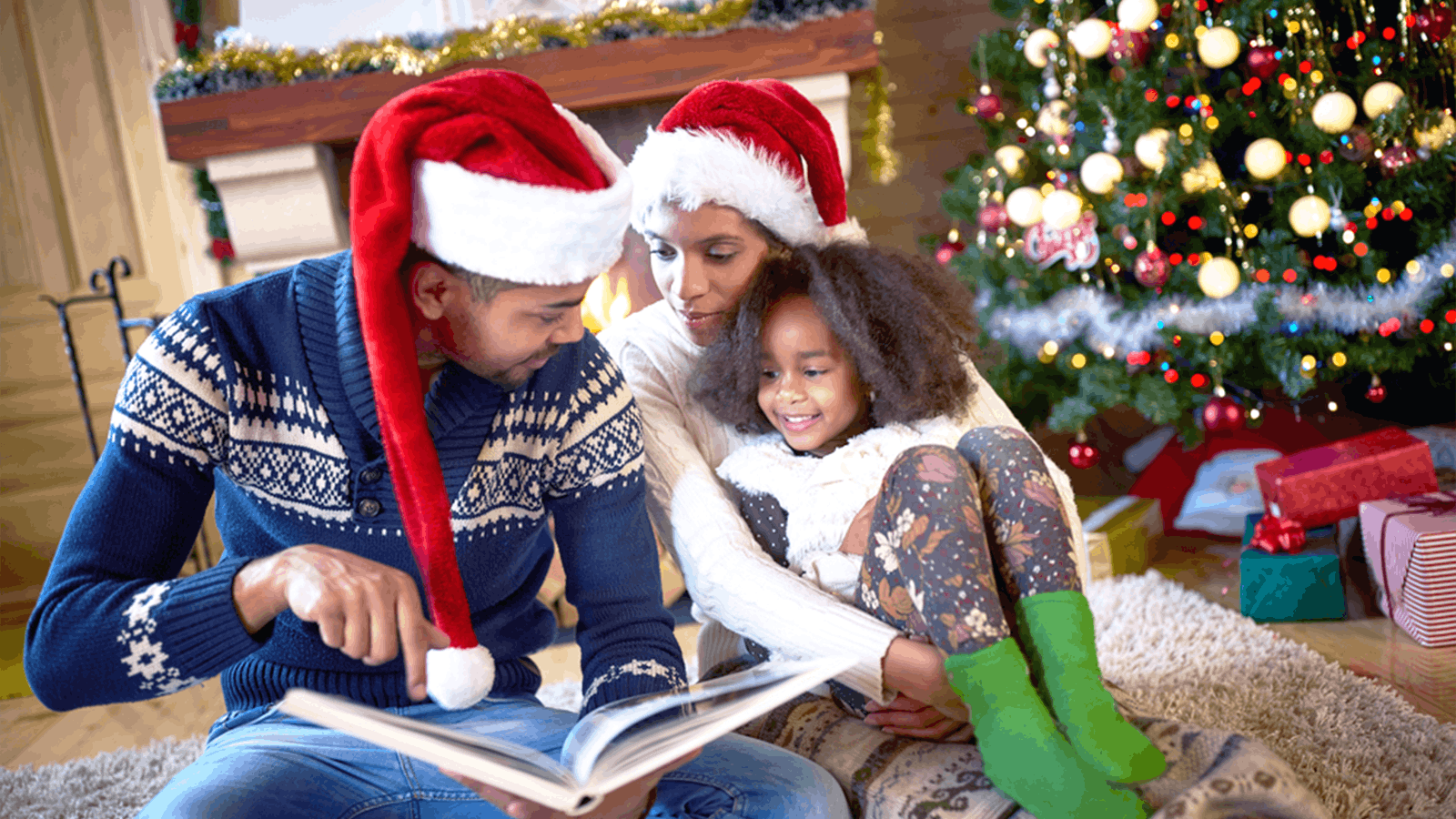 A family wearing Santa hats sits by a decorated Christmas tree, reading a book together. The parents smile and the child, sitting on her mothers lap, looks happy. Wrapped gifts are visible under the tree.