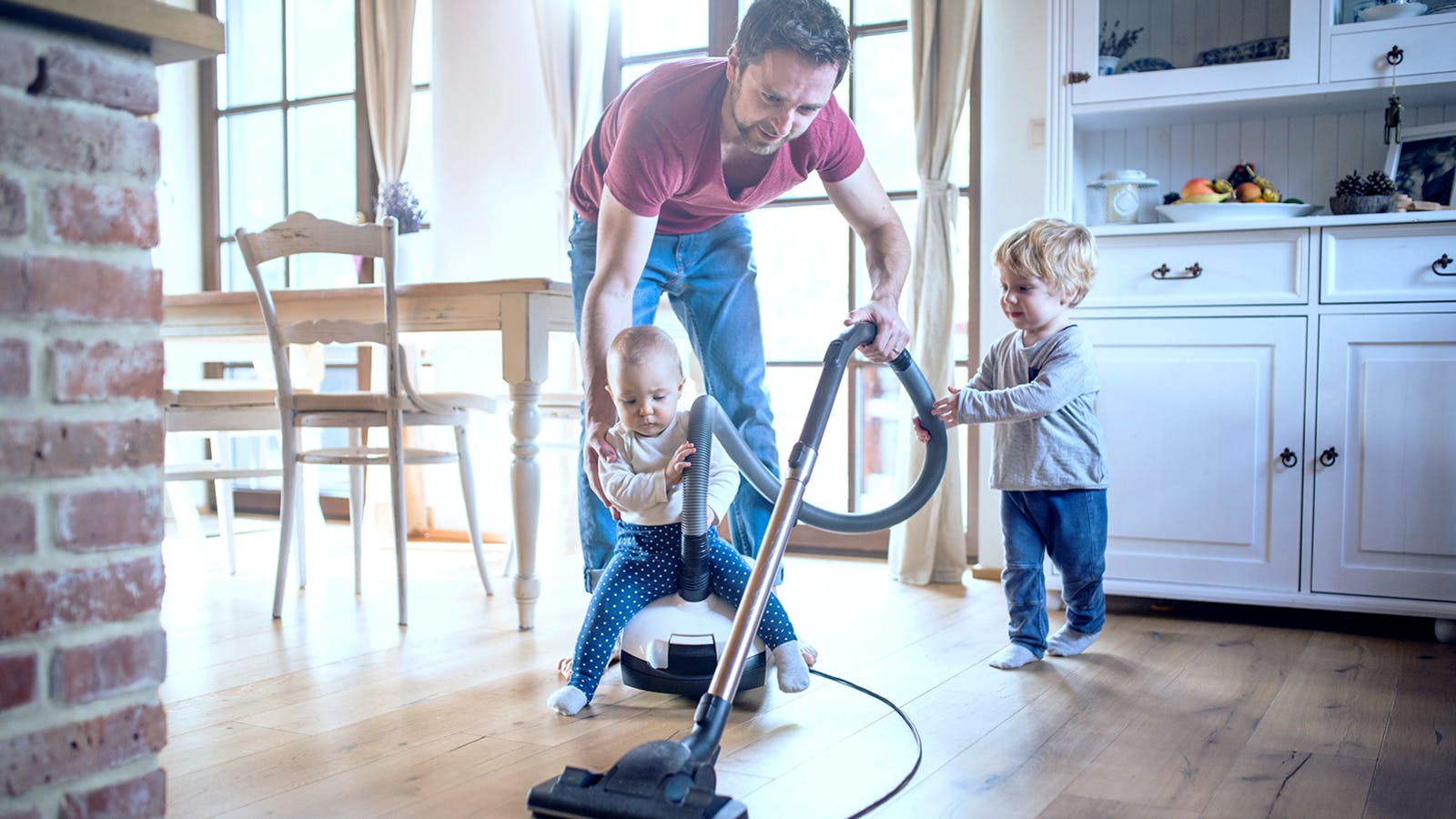 A man vacuums a wooden floor in a bright kitchen while a baby sits on the vacuum cleaner and a young child smiles and watches nearby.