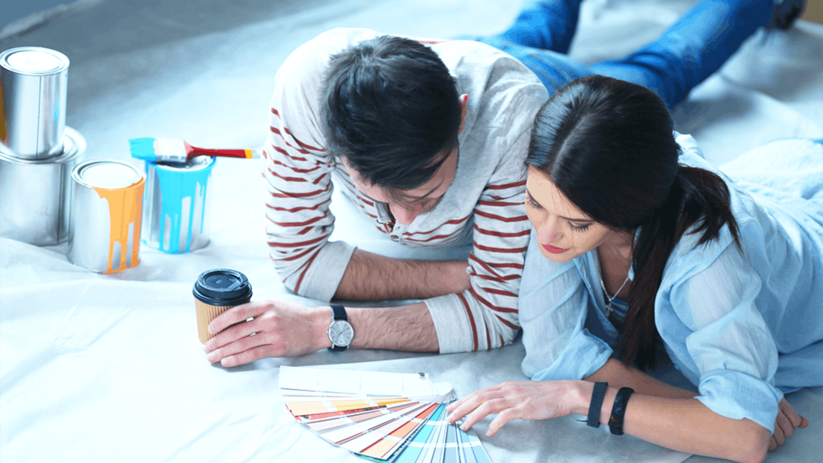 A man and woman lie on the floor looking at paint color swatches. Paint cans, brushes, and a coffee cup are nearby, suggesting they are planning to paint a room together.