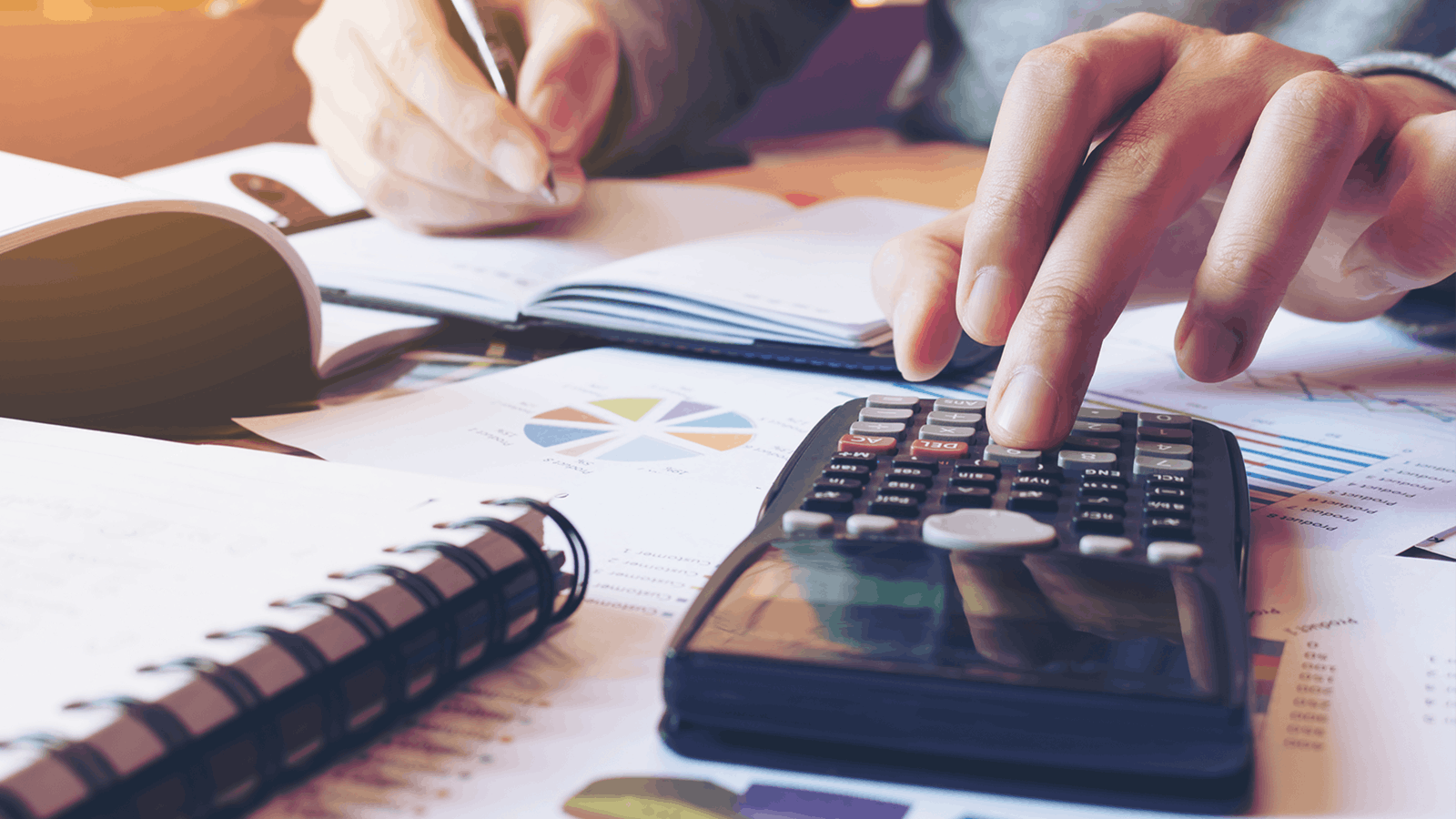 A person uses a calculator and writes in a notebook, surrounded by papers, charts, and a pair of glasses on a desk, suggesting financial planning or budgeting activities.