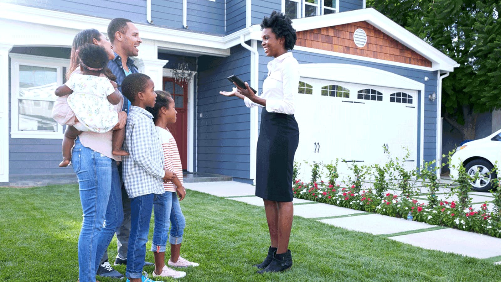 A smiling real estate agent talks with a family of five, including two adults and three children, standing on the lawn in front of a modern blue house with a garage and white trim.