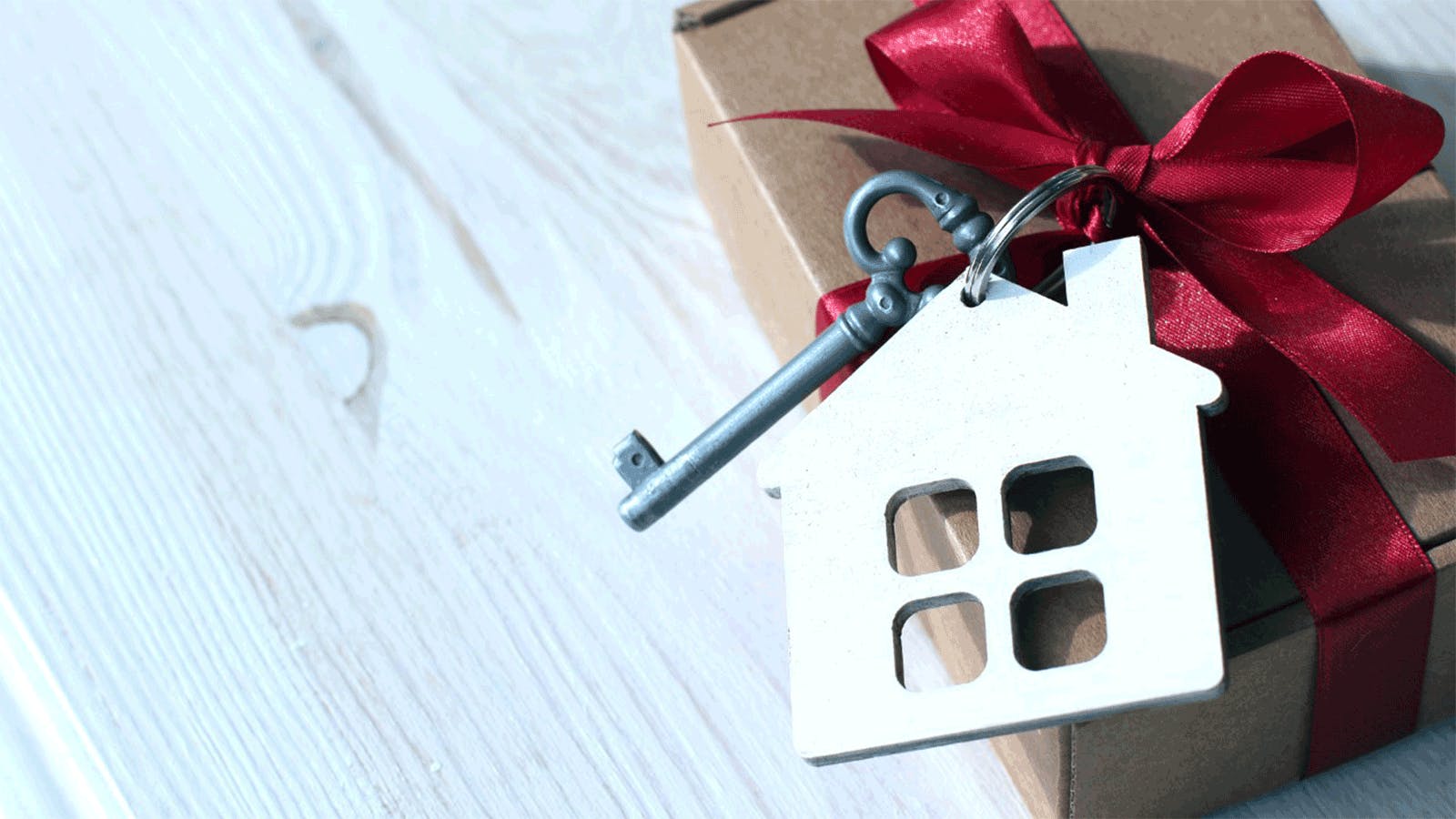 A gift box tied with a red ribbon sits on a white wooden surface, with a vintage-style key and a house-shaped keychain placed on top, symbolizing a new home or housewarming gift.