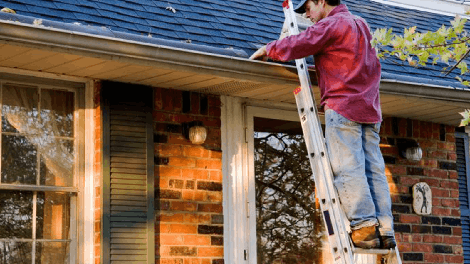 A person stands on a ladder cleaning leaves and debris from the gutter on the roof of a brick house with dark shutters and a covered porch.