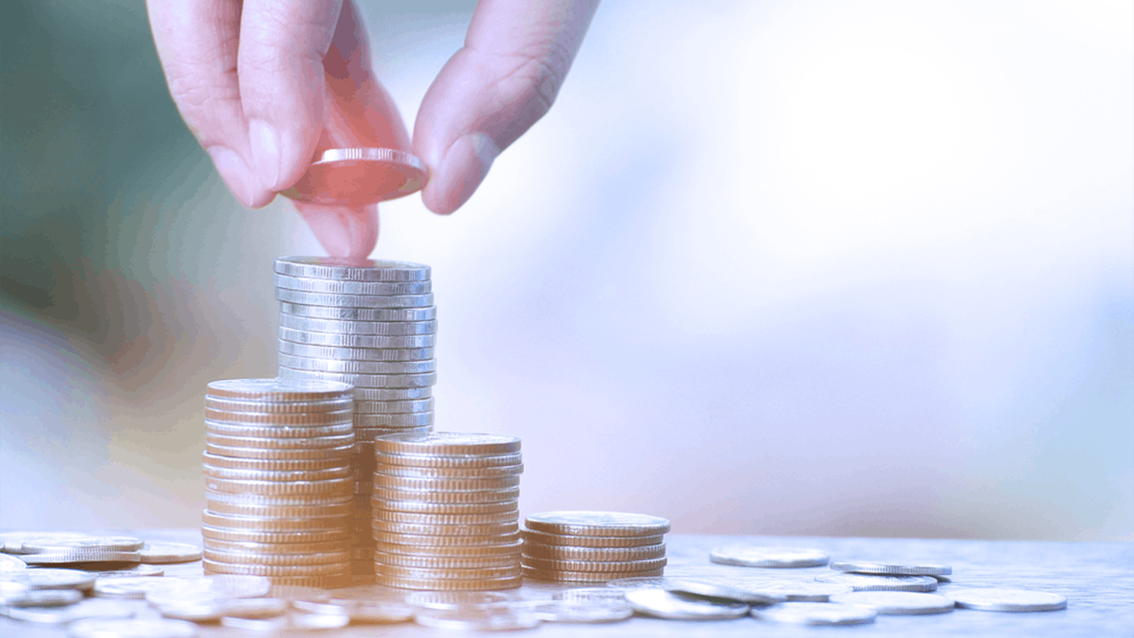 A hand places a coin on top of a stack of coins, with several other stacks and loose coins on a surface, symbolizing saving or financial growth.