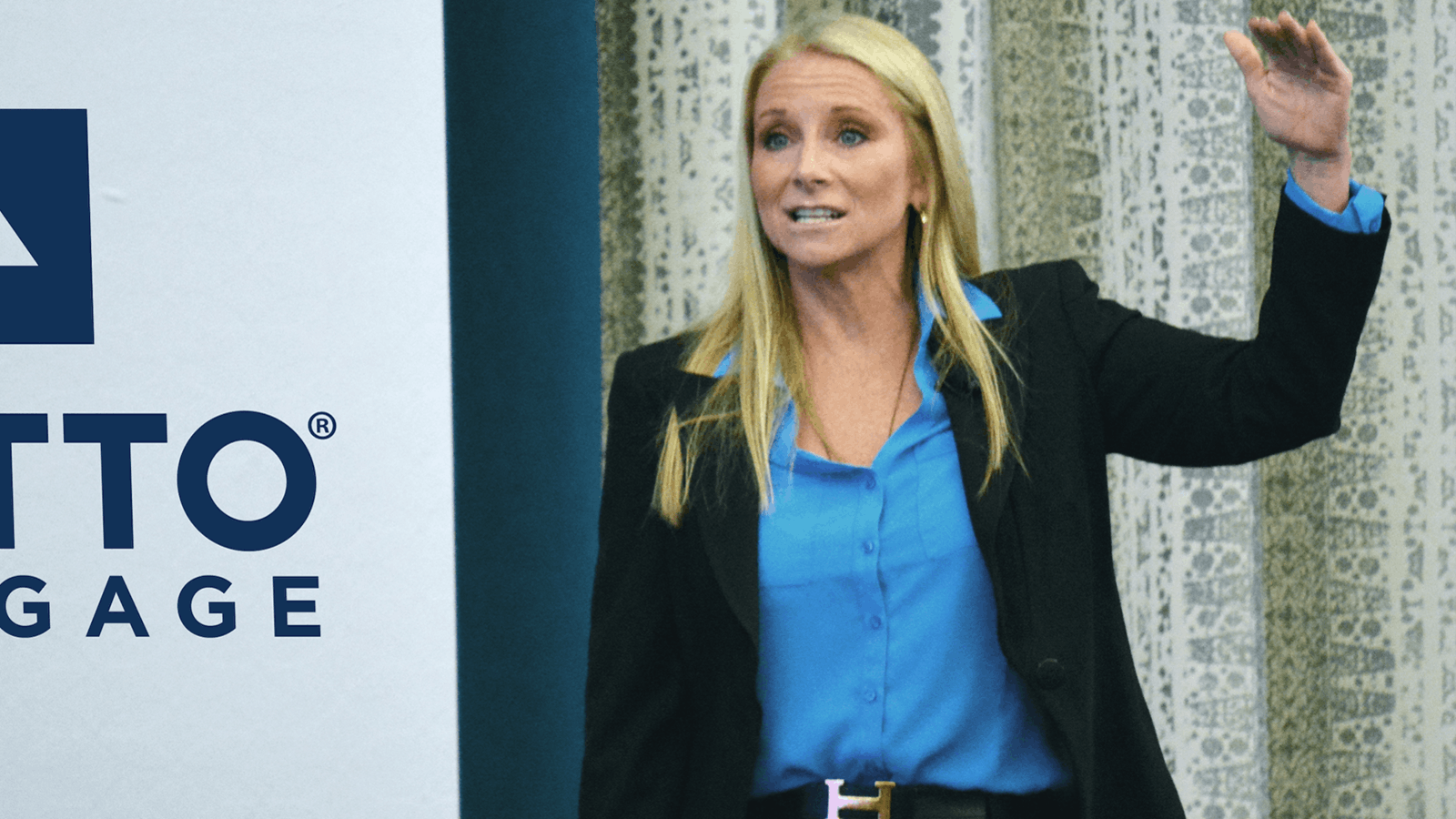 A woman in a blue shirt and black blazer stands speaking next to a banner with partial text reading OTTO MORTGAGE, holding one hand raised. Curtains are visible in the background.