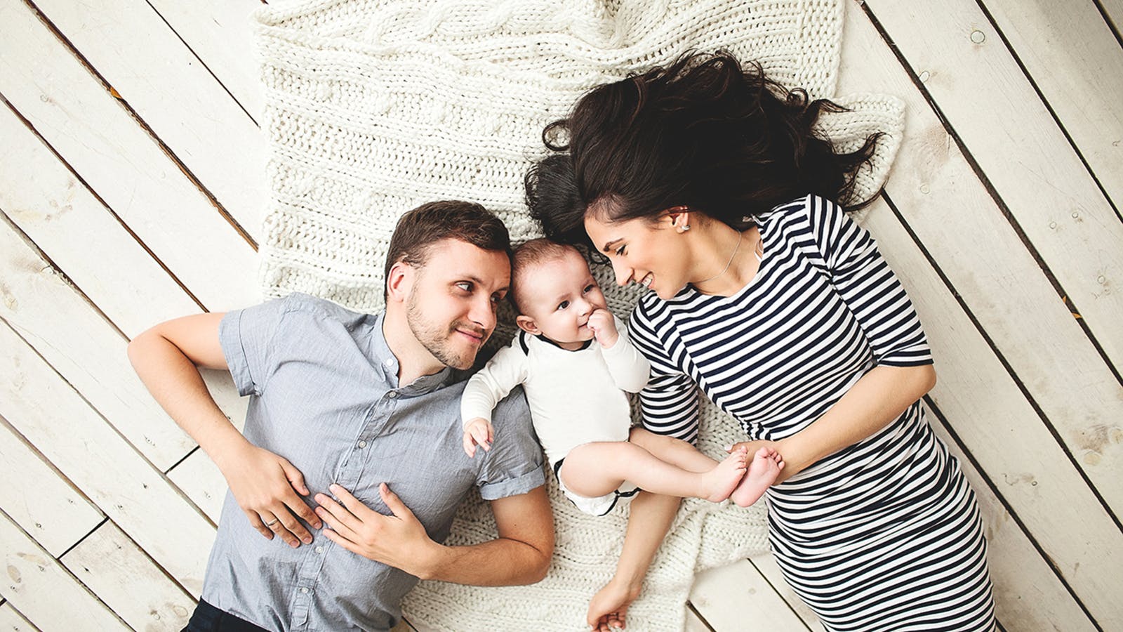 A man, woman, and baby lie on a cream-colored knitted blanket on a wooden floor. The man and woman face each other, smiling, while the baby looks up, holding the woman’s hand. The woman wears a striped dress.