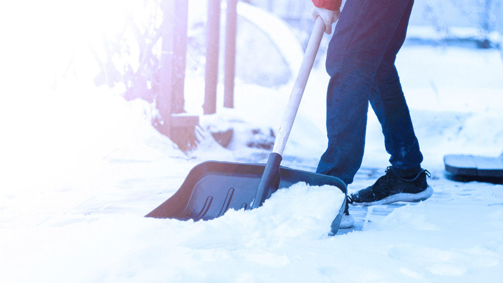Person shoveling snow from a sidewalk with a large snow shovel, wearing dark pants and black shoes, on a cold winter day. The background is bright and slightly out of focus.