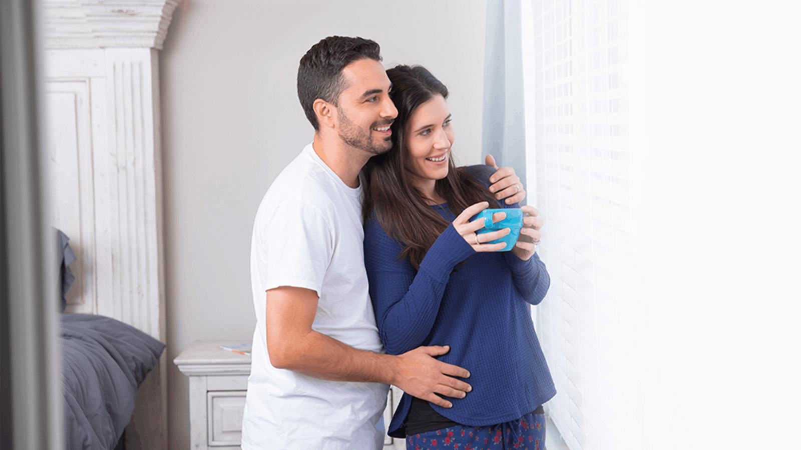 A smiling couple stands by a window; the man hugs the woman from behind as she holds a blue mug, both looking outside. The woman rests her hand on her belly, and they appear happy and relaxed.