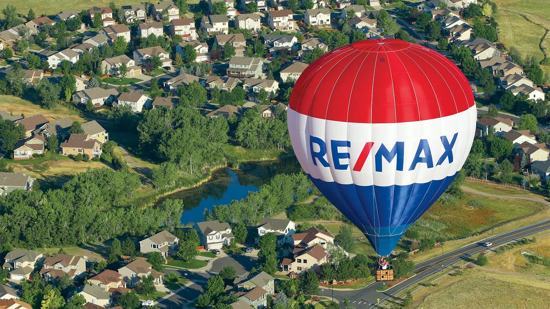 A RE/MAX hot air balloon floats above a suburban neighborhood with houses, trees, and a small pond visible below.