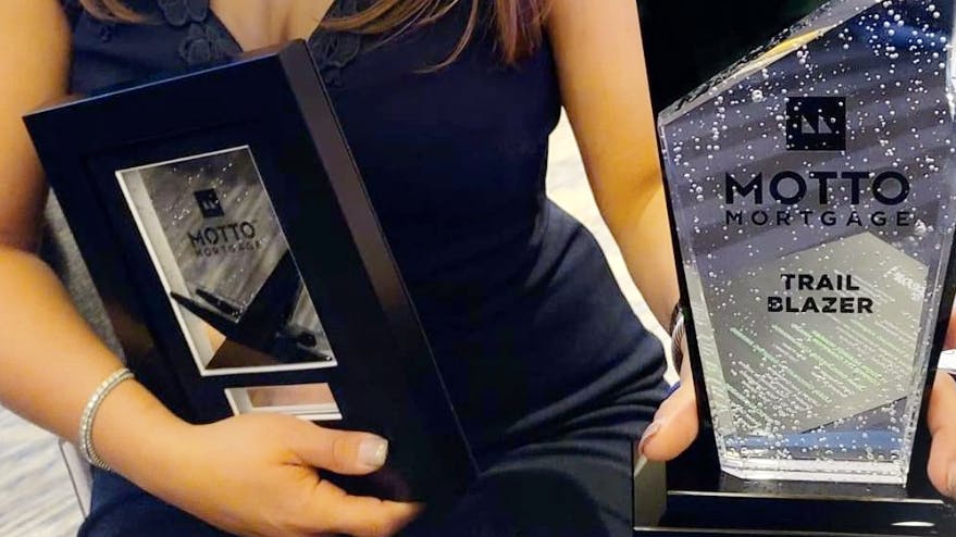 A person in a dark dress holds two Motto Mortgage awards, one labeled Trail Blazer, both featuring glass and black designs. Only the person’s torso and arms are visible.