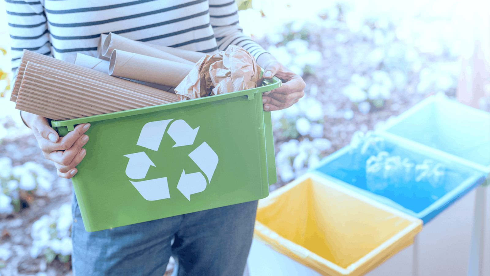 A person holding a green recycling bin filled with cardboard and paper, standing near blue, yellow, and white recycling bins in an outdoor setting.