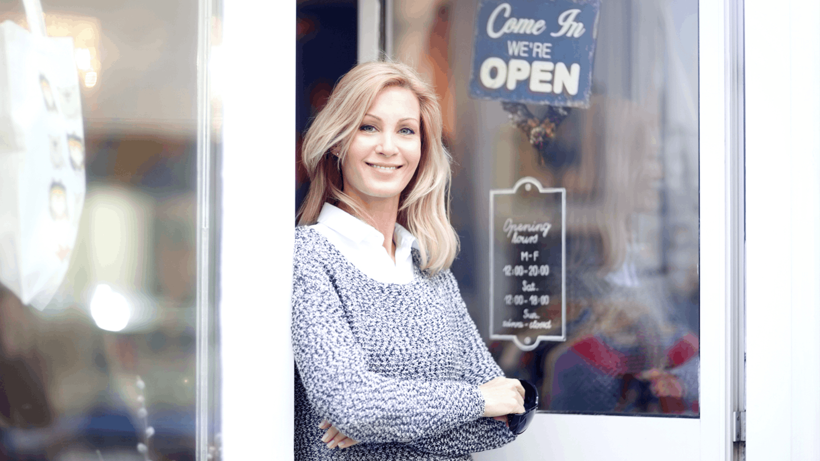 A smiling woman with blonde hair, wearing a gray sweater over a white collared shirt, stands in the doorway of a shop with a sign that says Come In, We’re OPEN. The shop hours are also posted on the door behind her.