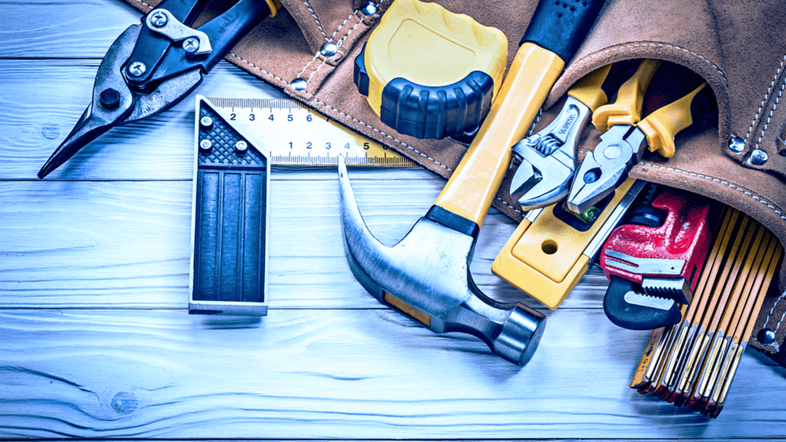 A set of various hand tools including a hammer, tape measure, pliers, and wrench laid out on a wooden surface next to a brown tool belt.