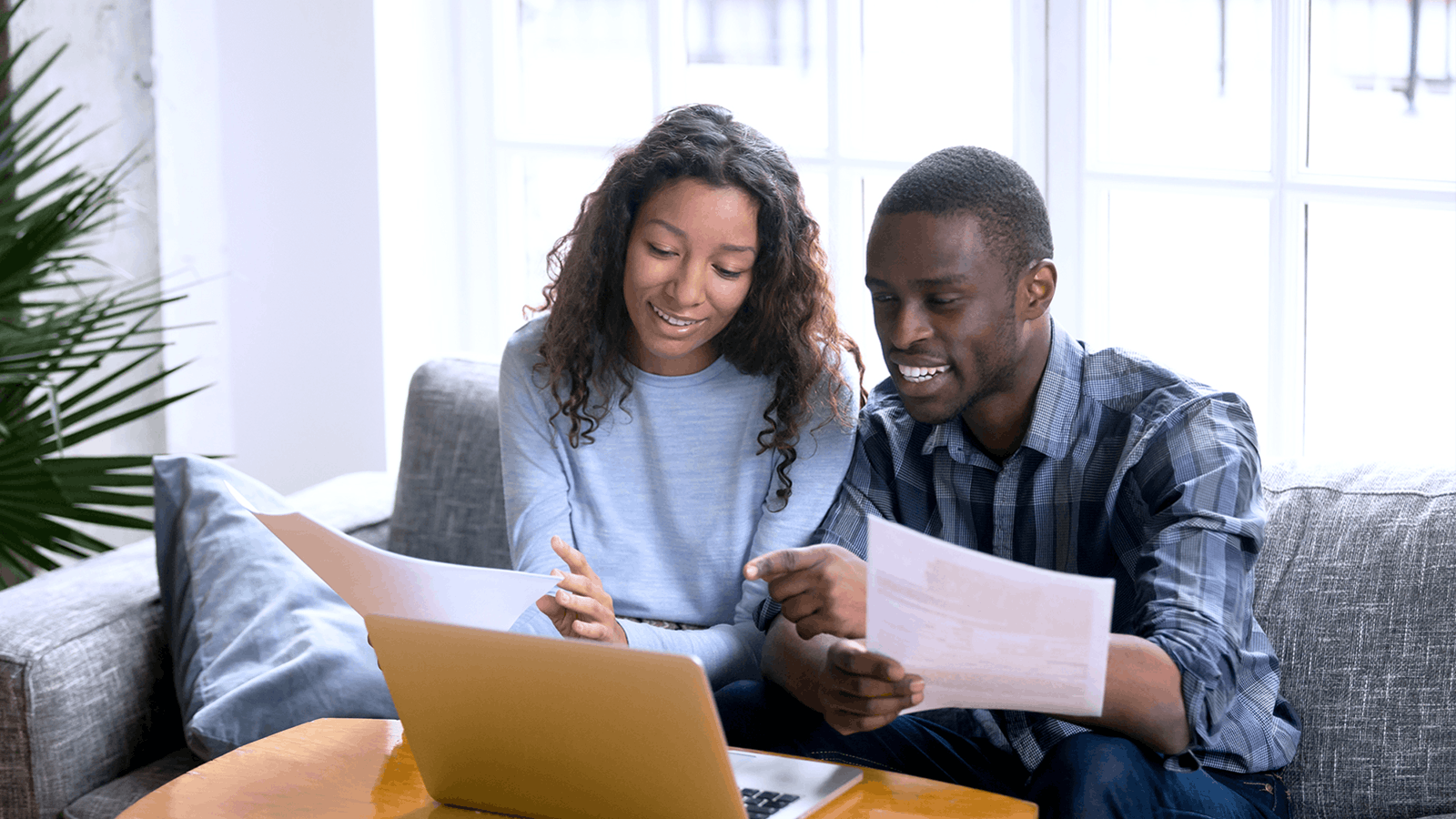 A smiling couple sits on a couch looking at papers and a laptop together, appearing to discuss or review documents in a bright, cozy living room.