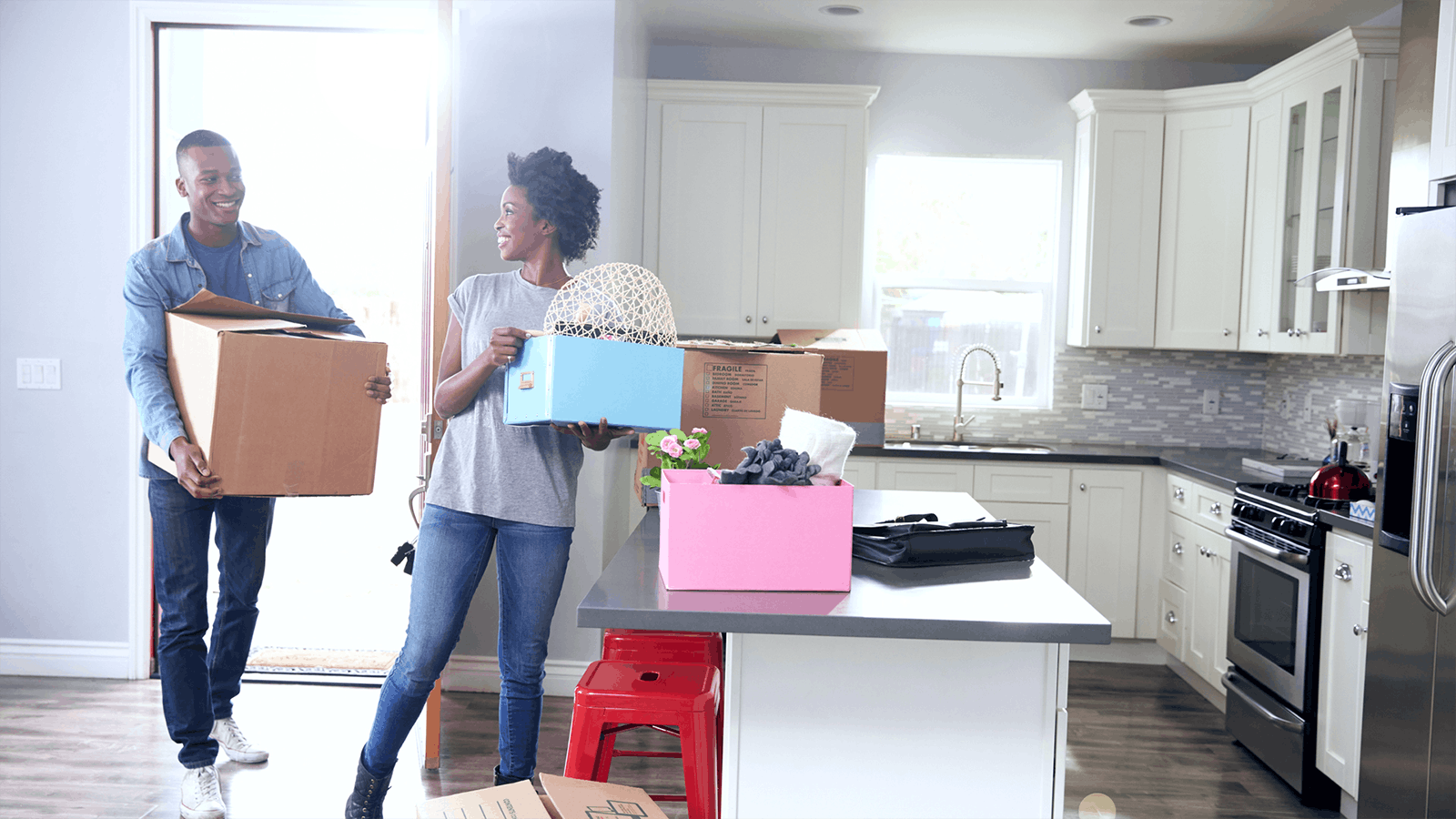 A smiling couple carries boxes into a bright, modern kitchen, unpacking and moving into their new home. Cardboard and colorful storage boxes are scattered on the island and floor.
