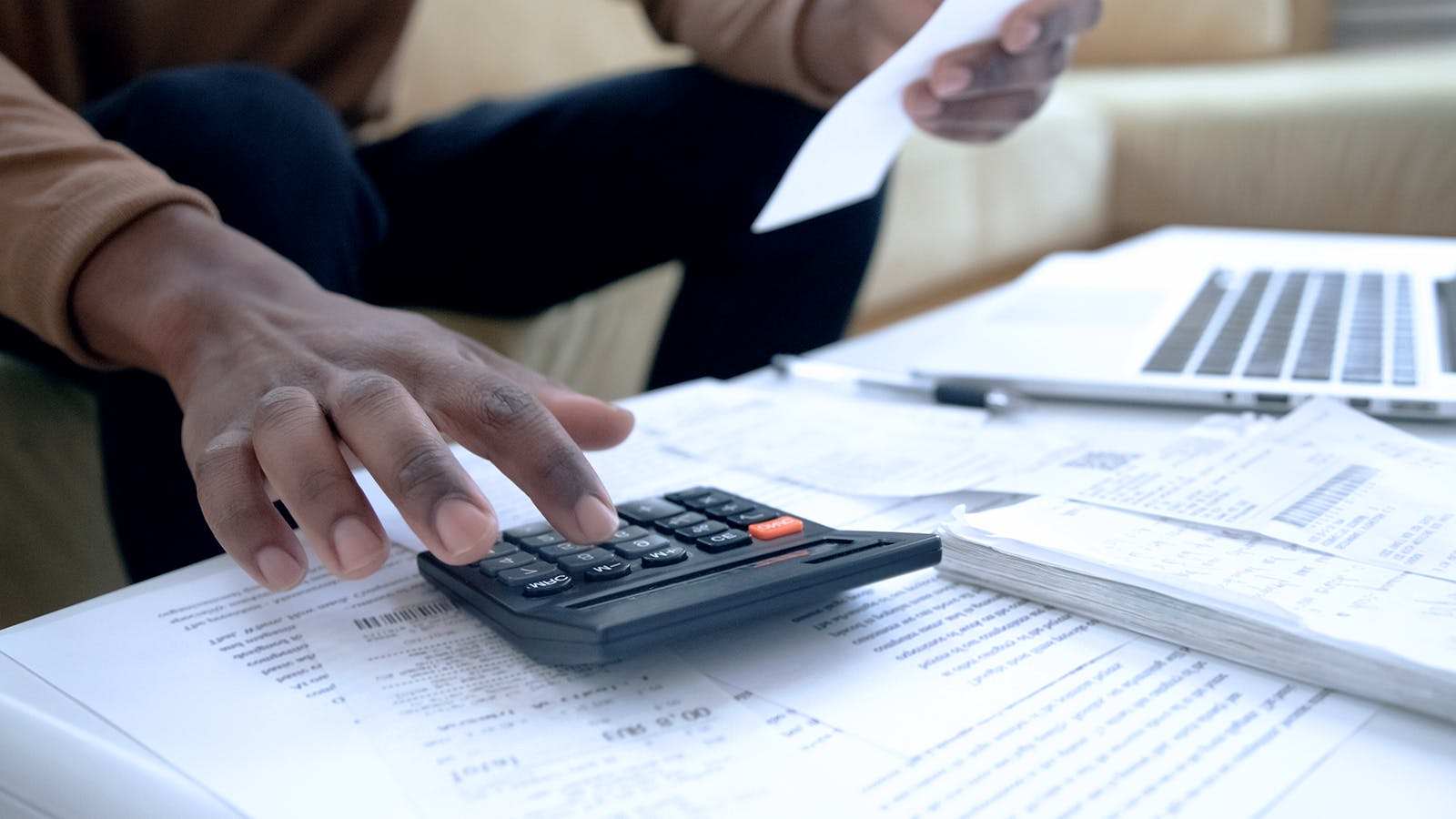 A person uses a calculator while holding a receipt. Papers, receipts, and a laptop are spread across the table, suggesting budgeting or bill management at home.