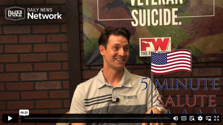A man in a gray polo shirt sits smiling during an interview. Behind him are signs reading VETERAN SUICIDE and DAILY NEWS Network. An American flag graphic and video player controls appear at the bottom.