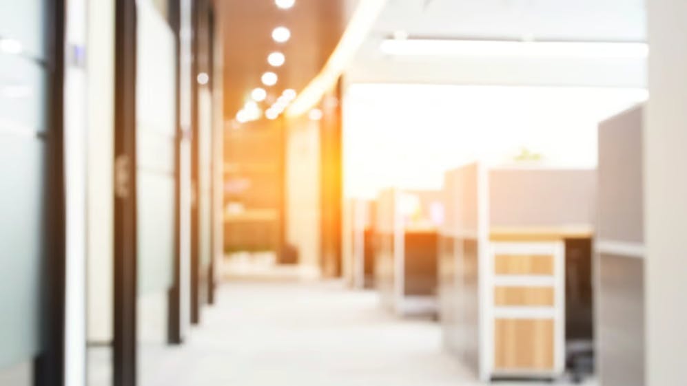 A blurred image of a modern office space with cubicles, bright lighting, and sunlight streaming in through windows, creating a warm and inviting atmosphere.