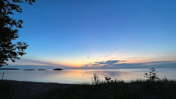 A calm lake at sunset with a clear sky fading from blue to orange, a few silhouetted trees and shrubs in the foreground, and small islands visible on the horizon.