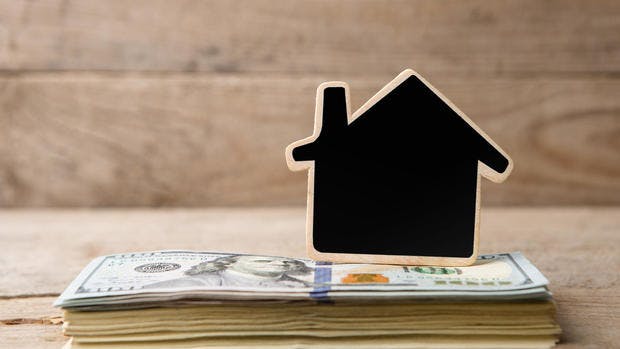 A small black house-shaped model sits on top of a stack of hundred-dollar bills, symbolizing real estate investment or home financing, with a wooden background.