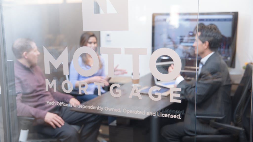 Three people sit at a conference table having a discussion in an office, viewed through a glass door with the words MOTTO MORTGAGE and additional text printed on it.