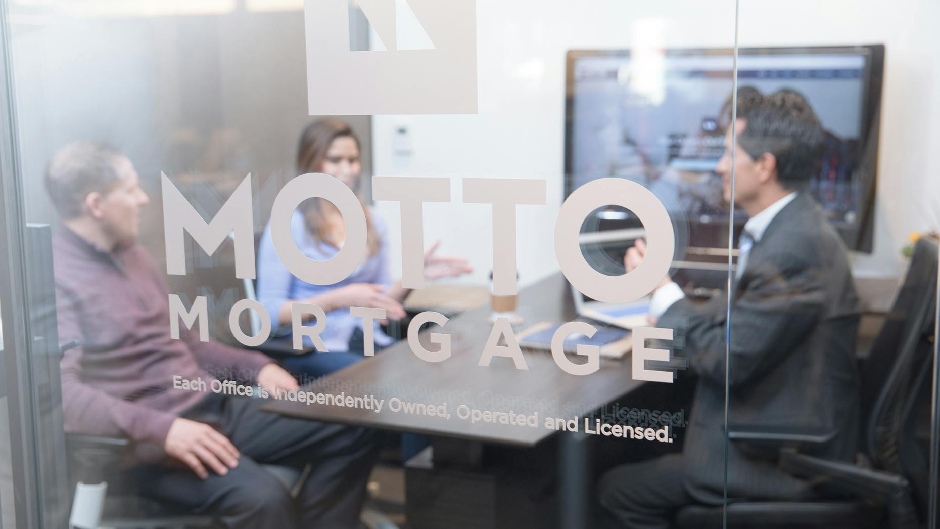Three people sitting at a desk behind a glass wall with the Motto Mortgage logo