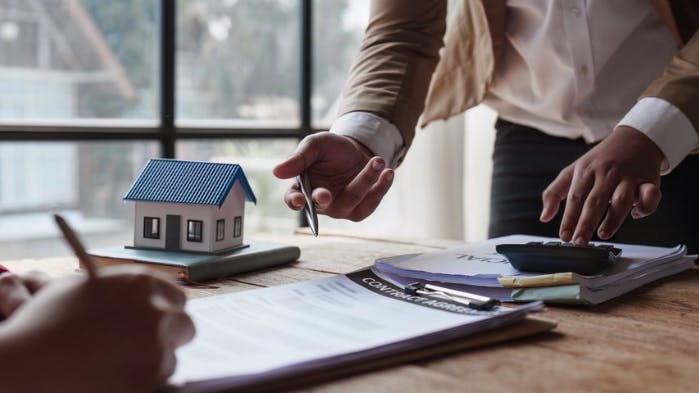 Two people meet at a table with paperwork, a calculator, and a small model house, suggesting a real estate or financial discussion. One person gestures with a pen while the other points at documents.