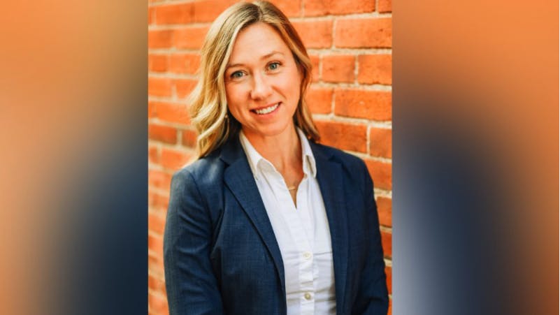 A woman with blonde hair, wearing a navy blazer and white shirt, stands smiling in front of a red brick wall.