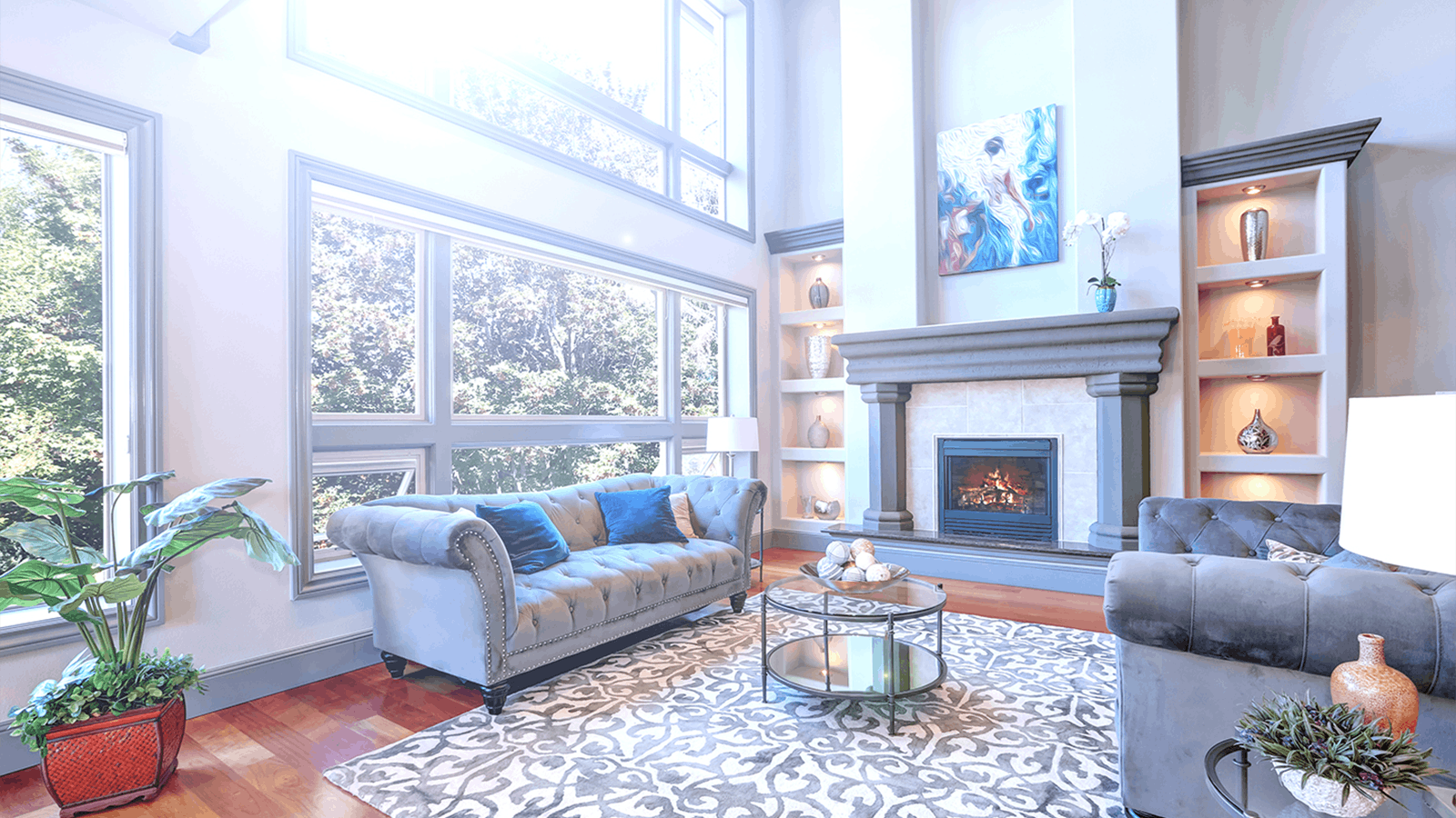 A bright, modern living room with large windows, blue velvet sofas, a patterned rug, a coffee table, and a decorative fireplace surrounded by built-in shelves with art and vases. Sunlight streams in, highlighting the airy space.