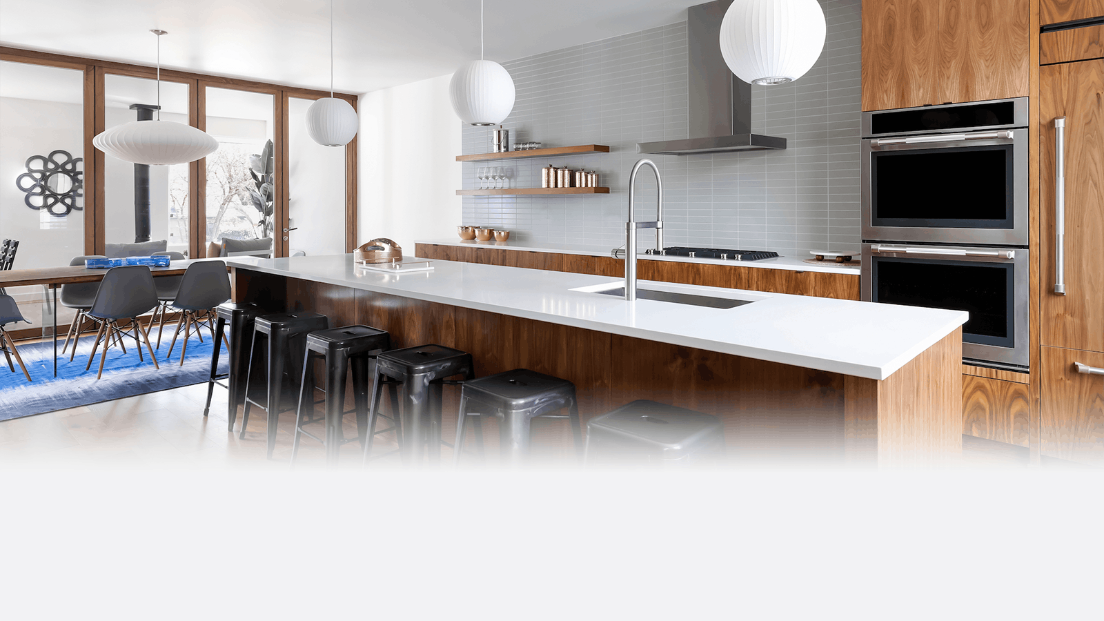 Modern kitchen with a long island, black stools, white countertops, wooden cabinets, built-in ovens, and pendant lights. Dining area with chairs and a blue rug is visible in the background.