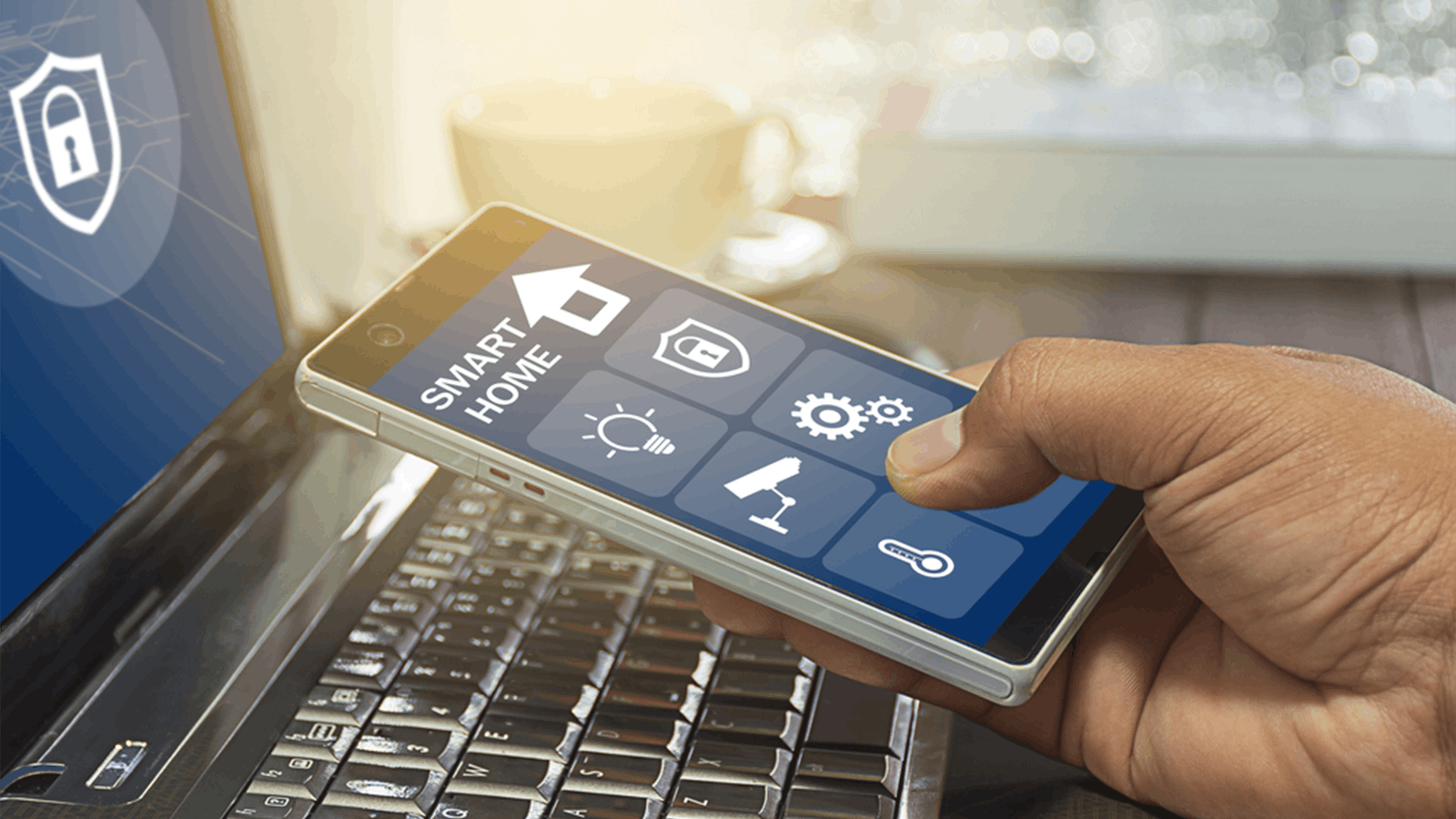 A hand holds a smartphone displaying smart home controls, including icons for lighting, security, and temperature, in front of an open laptop on a desk near a window with sunlight.