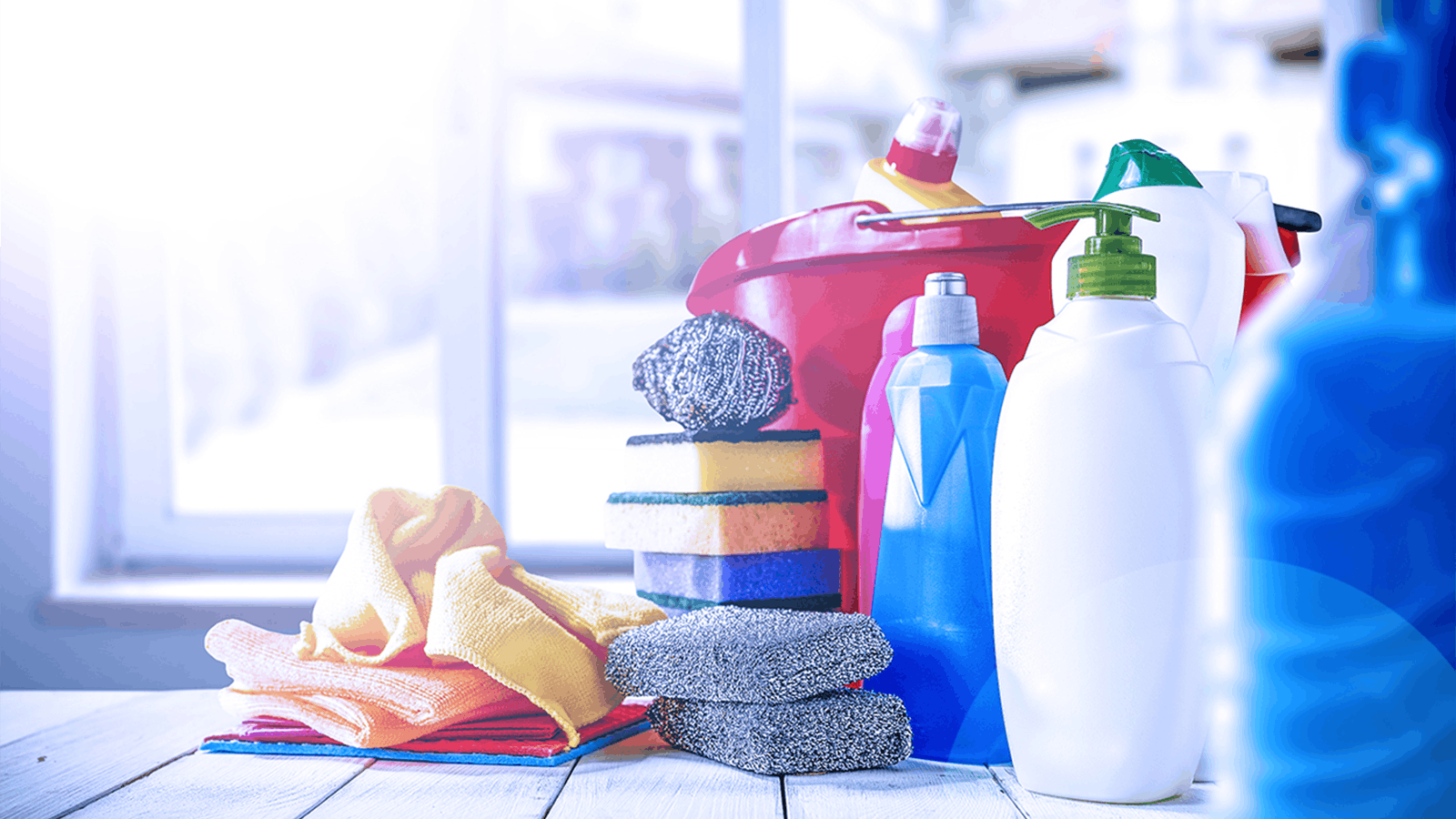 Assorted cleaning supplies, including bottles, sponges, steel wool, microfiber cloths, and a red bucket, are arranged on a white surface near a sunlit window.