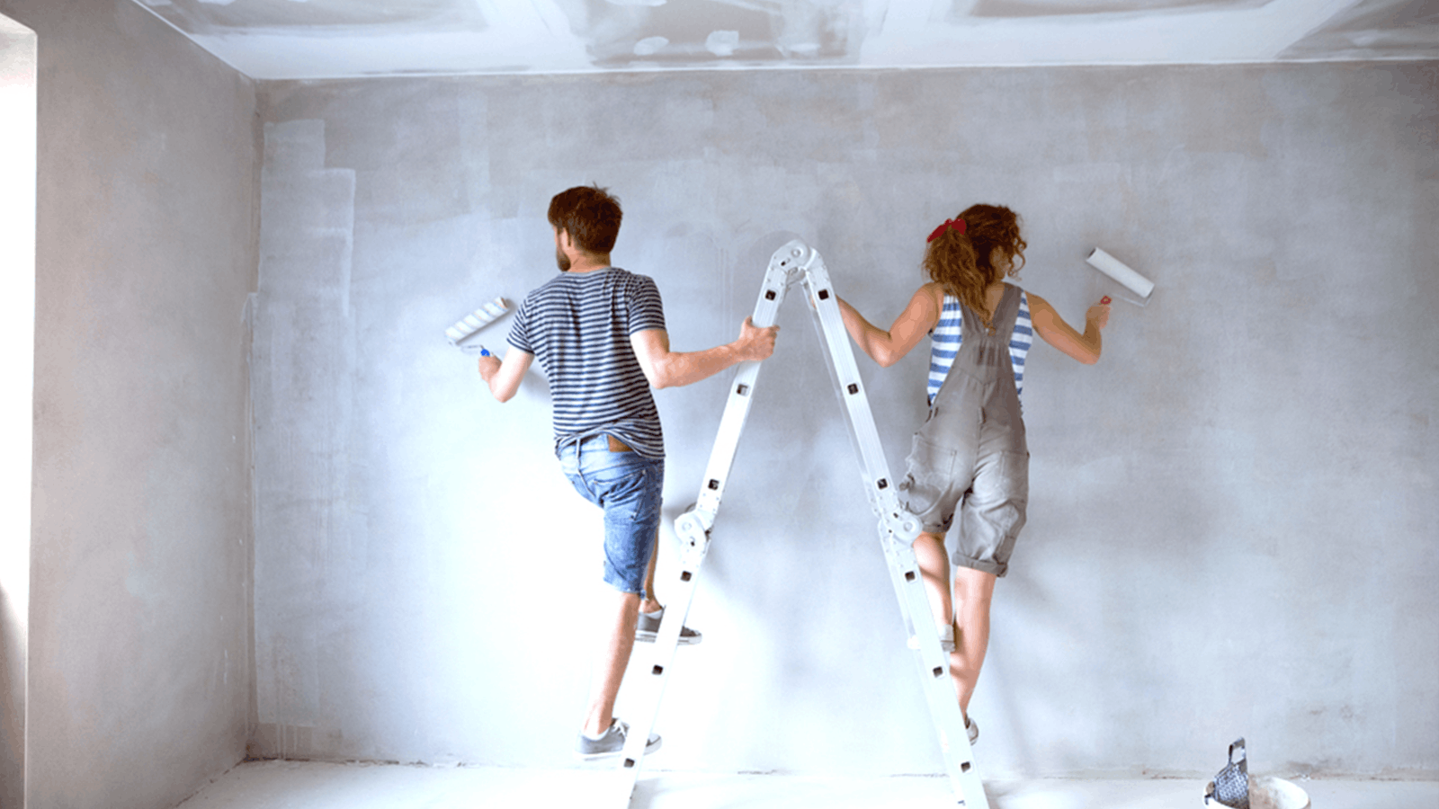Two people painting a wall white; one stands on a ladder while the other stands on the floor. Both are using paint rollers, wearing casual clothes, and working in a room under renovation.