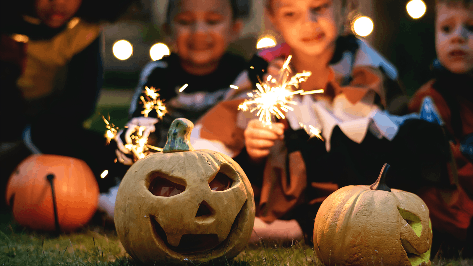 Children in costumes sit on grass holding sparklers, with two carved pumpkins in the foreground. The scene is festive and illuminated by string lights, suggesting a Halloween celebration.