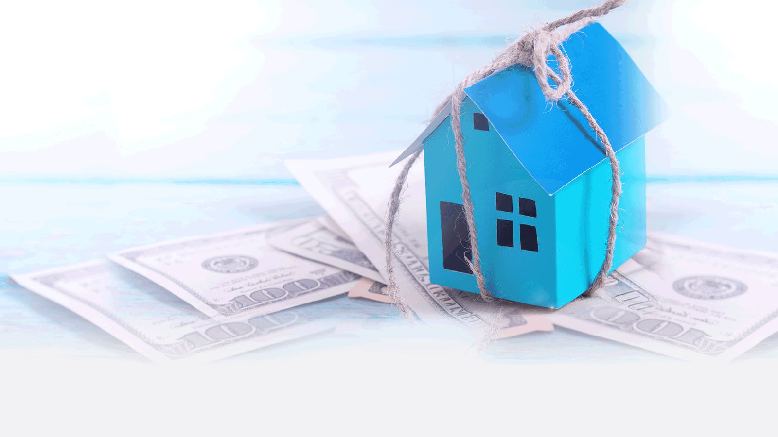 A small blue paper house tied with string sits on top of scattered U.S. hundred-dollar bills, symbolizing home expenses, mortgage, or financial investment in real estate.