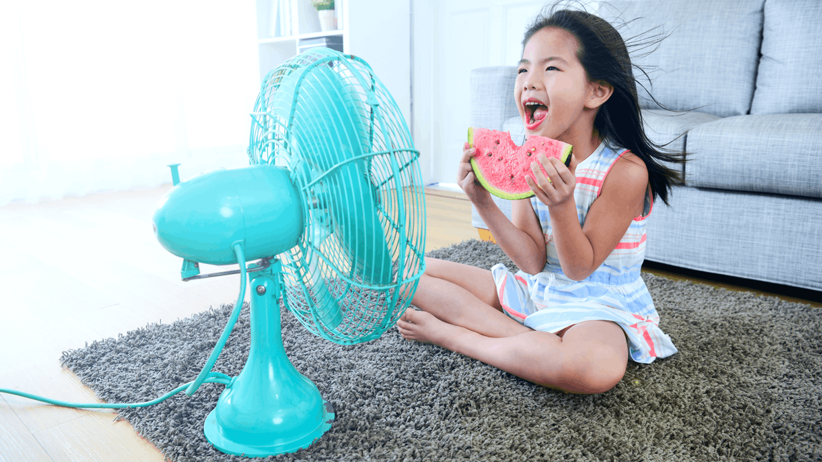 A young girl sits cross-legged on a rug indoors, eating a slice of watermelon and smiling as a turquoise electric fan blows air toward her. A grey sofa and bright window are in the background.