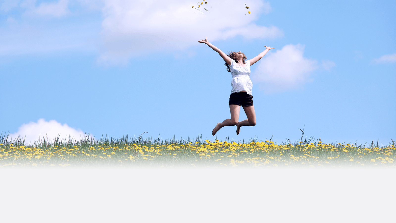 A woman in a white top and black shorts jumps joyfully in a grassy field with yellow flowers under a blue sky with scattered clouds. She has her arms raised as yellow petals float above her.